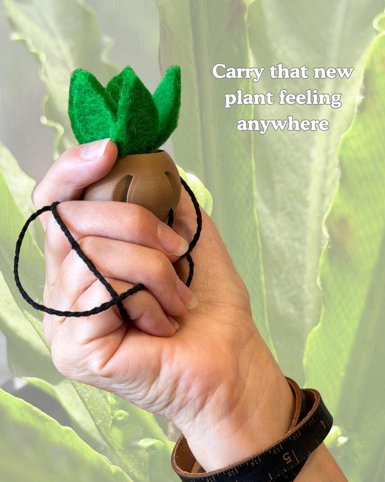 May include: A small brown pot with a vibrant green felt succulent plant attached to a thin black string. The text "Carry that new plant feeling anywhere" is printed in white on the right side of the object.