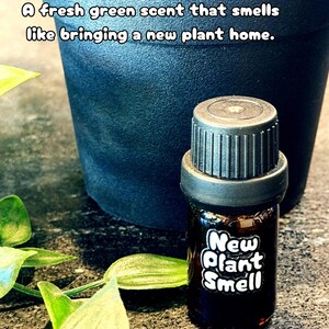 May include: A small amber glass bottle with a black ribbed cap and a white label featuring the text New Plant Smell. The bottle is positioned in front of a dark gray plant pot. The text A fresh green scent that smells like bringing a new plant home is visible at the top.