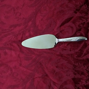 Op de afbeelding: Een zilveren taartschep met een decoratief bloemenontwerp op de steel. De schep ligt op een rode stoffen achtergrond.