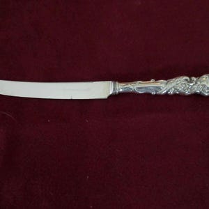 May include: A silver butter knife with a decorative handle. The blade is smooth and the handle features an ornate floral design. The knife is set against a dark red background.