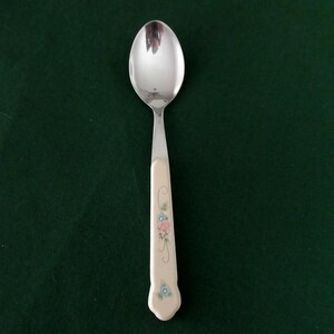 May include: A silver spoon with a floral design on the handle. The spoon has a shiny, reflective bowl and a cream-colored handle decorated with pink and blue flowers. The spoon is set against a dark green background.