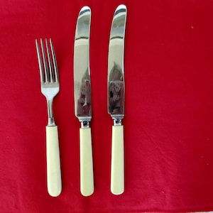 May include: A fork and two knives with cream-colored handles and silver blades. The silverware is arranged on a red surface. The blades of the knives are curved. The fork has four tines.