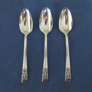 Puede incluir: Tres cucharas de plata con cuencos ovalados y mangos ornamentados. Los mangos tienen un diseño floral detallado. Las cucharas están dispuestas verticalmente sobre un fondo azul oscuro.