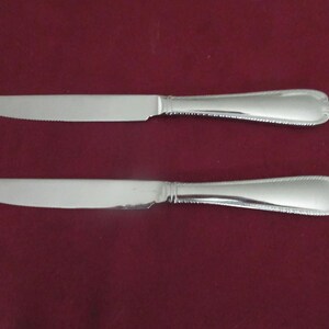 May include: Two silver-toned dinner knives with serrated blades and smooth, polished handles. Each handle features a subtle beaded or rope-like decorative border along the edges, terminating in a rounded, slightly flared end design.