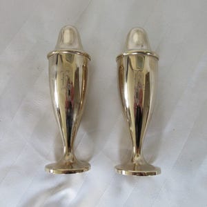 May include: Two matching, shiny, gold-colored salt and pepper shakers. Each shaker has a rounded top and a slender, elongated body that flares out slightly at the base. The shakers are engraved with a letter. The shakers are on a white surface.
