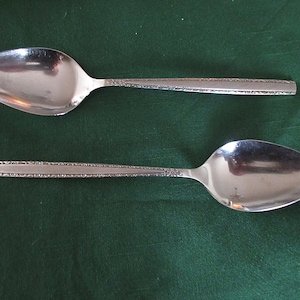 May include: Two silver spoons with an ornate design on the handle. The spoons are on a green surface.