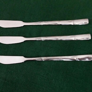 Könnte beinhalten: Drei silberne Buttermesser mit Bambus-Griffen liegen auf einer dunkelgrünen Oberfläche. Die Messer haben eine gebogene Klinge und eine polierte Oberfläche. Die Griffe weisen ein strukturiertes Design auf.