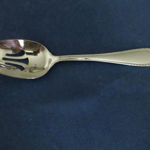 May include: A silver-toned slotted serving spoon with a polished finish. The bowl of the spoon features three distinct cutouts, and the handle is designed with a decorative beaded or rope-like border detail along its entire length.