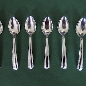 May include: Six silver spoons arranged in a row on a dark green surface. The spoons have a classic design with oval bowls and detailed handles. The spoons are reflective, catching the light.