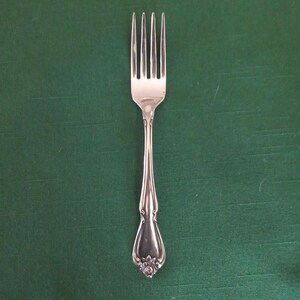 Può includere: Una forchetta d'argento a quattro rebbi, su sfondo verde. Il manico presenta un motivo floreale decorativo alla base. Questa posata è probabilmente per la ristorazione.