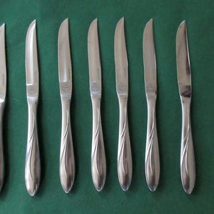 May include: A collection of seven stainless steel butter knives with a polished finish and curved handles. The blades are slightly curved. The knives are displayed on a dark green surface, offering a classic and elegant aesthetic for dining.
