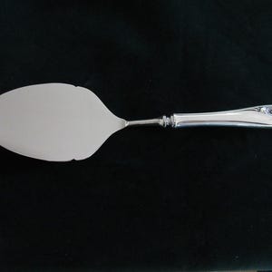 Puede incluir: Una espátula plateada para pasteles con una hoja ovalada y un mango decorativo. El mango presenta una serie de pequeños adornos transparentes cerca del extremo. Diseñado para servir pasteles y repostería.