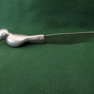Può includere: Un coltello da burro color argento con un manico a forma di uccello. La testa dell'uccello è nella parte superiore del manico e la lama si estende dal corpo dell'uccello. Il coltello è su una superficie verde.