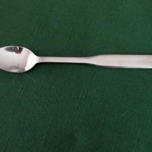 May include: A stainless steel spoon with a rounded bowl and a flat handle. The spoon is set against a dark green background, creating a contrast that highlights its polished surface. This utensil is suitable for serving or eating.