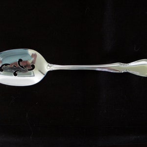 May include: A silver slotted serving spoon with an oval bowl and a decorative handle. The bowl has three decorative cut-out designs. The spoon is made of a shiny, reflective metal, likely stainless steel.