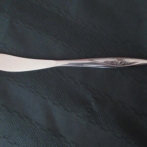 May include: A silver butter knife with a rounded blade and a floral pattern near the handle. The knife is made of shiny metal and is designed for spreading butter or other soft spreads. The knife is approximately 18 cm long.