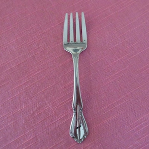 Peut inclure: Une fourchette argentée à quatre dents, posée sur une surface rose texturée. La fourchette a un manche décoratif avec un design détaillé. Vue de dessus de la coutellerie.