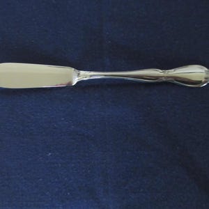 Può includere: Un coltello da burro argentato con una lama arrotondata e un manico decorativo. L'impugnatura ha una forma bulbosa con dettagli ornati. Il coltello è appoggiato su uno sfondo blu scuro.