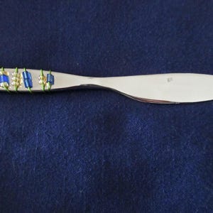 May include: A silver butter knife with a decorative handle. The handle is adorned with blue and gold beads wrapped with green wire. The knife rests on a dark blue surface.