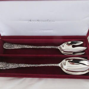 May include: A silver serving spoon and fork set with floral detailing, presented in a burgundy velvet-lined box. The set is from the "American Silverman's Collection". Ideal for serving salads or other dishes.