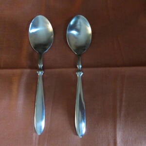 May include: Two matching silver-toned metal spoons with smooth, rounded bowls and slender, tapered handles. The spoons feature a polished, reflective finish and a simple, clean design with a slight decorative transition between the bowl and handle.