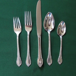 Puede incluir: Un juego de cinco cubiertos plateados, que incluye un cuchillo, dos tenedores y dos cucharas. Los cubiertos tienen diseños ornamentados y están dispuestos sobre una superficie verde.