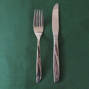 Puede incluir: Un juego de tenedor y cuchillo de acero inoxidable sobre un fondo verde. El tenedor tiene cuatro púas y el cuchillo tiene una hoja curva. Ambos utensilios tienen un diseño de estrella en la base del mango. Vajilla.