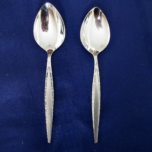 May include: Two silver spoons with a reflective surface and a decorative handle. The spoons have a floral design near the bowl and a star-like emblem. The spoons are set against a dark blue background.