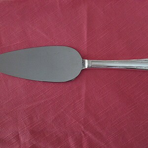 May include: A silver cake server with a wide, oval blade and a decorative handle. The server is made of shiny metal and is set against a red background.