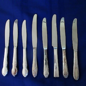 May include: A set of eight silver-toned metal dinner knives arranged in a row. Each knife features a smooth, polished blade and a handle with varying decorative embossed patterns, ranging from simple curved lines to intricate floral motifs.