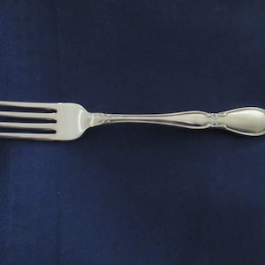 Könnte beinhalten: Eine silberne Gabel mit vier Zinken und einem dekorativen Griff. Der Griff hat ein verziertes Blumendesign am Ende und an den Rändern. Die Gabel ist vor einem dunkelblauen Hintergrund platziert.
