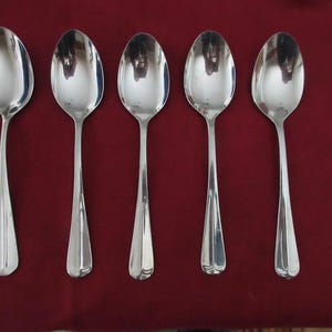 Puede incluir: Cinco cucharas plateadas brillantes dispuestas en fila. Cada cuchara tiene un cuenco redondeado y un mango largo con un diseño decorativo acanalado cerca de la base. El conjunto se presenta sobre un fondo rojo oscuro.
