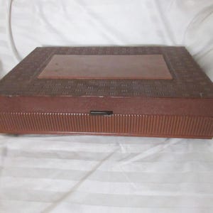 May include: A rectangular brown box with a textured lid and a rectangular inset. The box has a ribbed design on the sides and a small black latch. The box is sitting on a white surface.