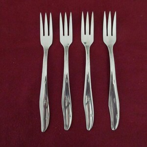 May include: Four silver forks with three tines each, set against a deep red backdrop. The handles feature a curved design with a starburst pattern. The forks are made of a shiny, reflective metal, suitable for dining.