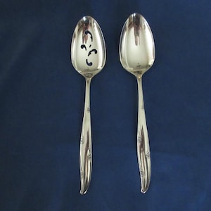 May include: Two silver serving spoons. One spoon has a decorative pattern on the bowl. Both spoons have a floral design on the handles. The spoons are set against a dark blue background.