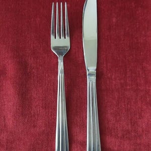 May include: A stainless steel fork and knife set against a red background. The fork has four tines and a long, ridged handle. The knife has a curved blade and a matching ridged handle. The silverware is clean and reflective.