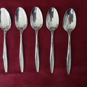 Pode incluir: Cinco colheres de prata com desenhos florais ornamentados nas alças. As colheres estão dispostas em uma fileira sobre uma superfície vermelha.