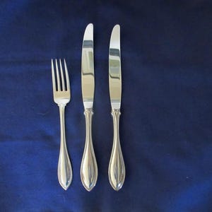 May include: A set of three pieces of polished silver-toned flatware consisting of one fork and two knives. Each piece features a smooth, tapered handle with a subtle central ridge design, reflecting light on the metallic surfaces.