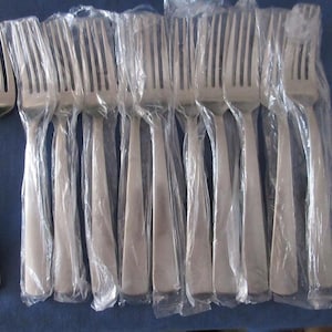 May include: A set of stainless steel forks, some individually wrapped in clear plastic. The forks have a simple, modern design with a smooth, elongated handle and four tines. The forks are arranged on a blue surface.