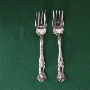 May include: Two silver forks with ornate handles and tines, set against a dark green background. The handles feature a detailed floral and vine design. The forks are likely vintage or antique.
