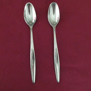 May include: Two shiny, silver spoons with oval bowls and slender handles. The handles feature a decorative etched design. The spoons are set against a deep red background, creating a contrast that highlights their polished surfaces. Ideal for serving.