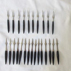 Könnte beinhalten: Eine Sammlung von Vintage-Serviergabeln und Vorspeisengabeln. Die Gabeln haben eine Zinke aus Edelstahl und einen schwarzen Griff. Die Vorspeisengabeln haben ein speerartiges Design. Die Gabeln sind auf einer weißen Oberfläche angeordnet.