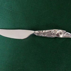 May include: A silver-toned metal butter knife featuring a smooth, curved blade and a handle intricately embossed with a stylized bird design. The handle depicts a detailed avian head with prominent eye and beak features in a polished finish.