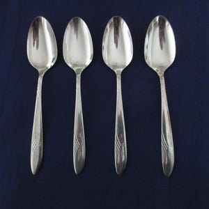 Könnte beinhalten: Vier silberne Löffel in einer Reihe angeordnet, jeder mit einer abgerundeten Schüssel und einem langen, schlanken Griff. Die Griffe weisen ein dekoratives, geätztes Design auf. Die Löffel sind vor einem dunkelblauen Hintergrund platziert.