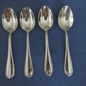 May include: A set of four polished silver-tone metal spoons arranged vertically. Each spoon features a smooth, rounded bowl and a slender handle decorated with a delicate beaded border along the edges and a small, subtle floral motif at the base.