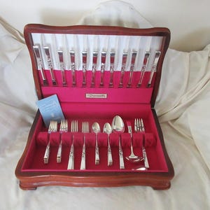 May include: A wooden flatware chest with a hinged lid, lined in bright red fabric. The interior features slots for silver-toned utensils, including forks, spoons, and a ladle. A small blue informational card is tucked inside the case.