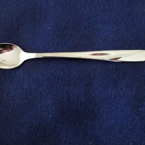 May include: A shiny, silver spoon with a rounded bowl and a slender, slightly curved handle. The spoon is made of a reflective metal, and it is set against a dark blue background.