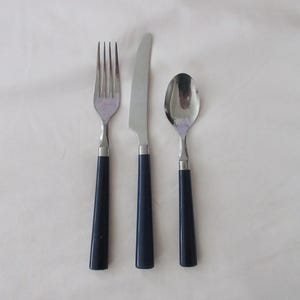 Puede incluir: Un juego de cubiertos de acero inoxidable que incluye un tenedor, un cuchillo y una cuchara. Cada pieza tiene un mango azul oscuro. El tenedor tiene cuatro púas y el cuchillo tiene una hoja redondeada.