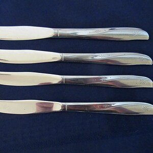 May include: Four butter knives with shiny, silver blades and handles. The handles have a decorative, curved design with a starburst pattern. The knives are arranged on a dark blue surface.