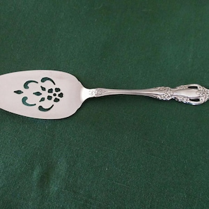 Puede incluir: Servidor de pastel plateado con un mango decorativo y una hoja ranurada con un diseño floral. El servidor está hecho de un metal brillante y está colocado sobre un fondo verde oscuro. Un utensilio de cocina para servir postres.